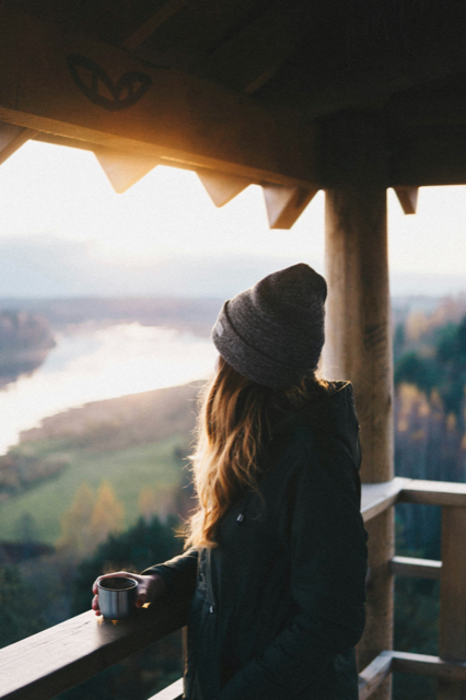 A young woman standing on a hilltop, gazing out at a beautiful panoramic view of mountains and a serene lake below, with the sun setting in the background, casting a warm golden glow over the landscape.